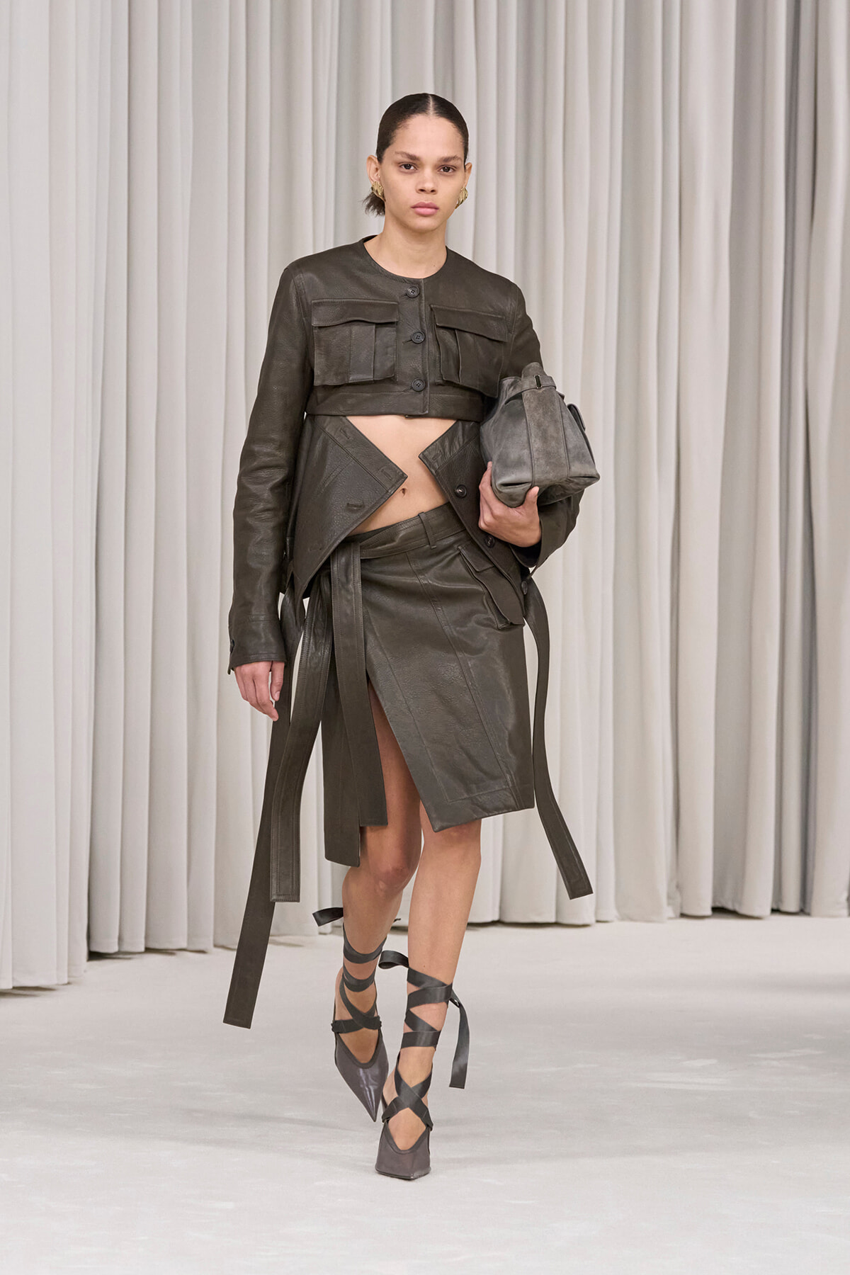 Model in a dark olive leather cropped jacket and asymmetrical wrap skirt with long belt straps, carrying a gray bag, and wearing crisscross ankle-strap heels on a runway.