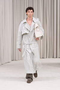 Male model walking a fashion runway in a light-gray crinkled trench coat with belt, brown wide-leg pants, brown loafers, and a white handbag.