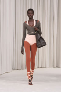 Model walking a runway in an olive wrap-top over a pale peach bodysuit, rust tights, pink wrap sandals, and a large black tote over the shoulder.