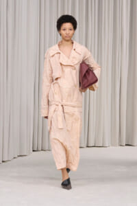 Model walking in a crinkled peach trench coat, carrying a burgundy handbag, in front of light curtains.