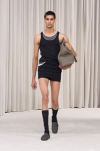 Male model walking a runway in a black sleeveless top and black shorts, carrying a gray tote bag in his right hand.