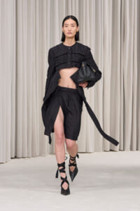 Model walking a runway in an all-black ensemble: cropped jacket, high-slit shorts/skirt, strappy heels, and a black handbag against a neutral backdrop.