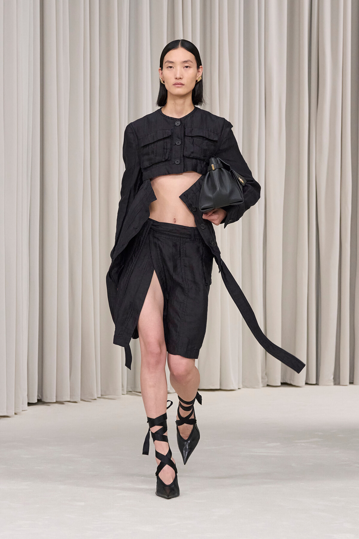 Model walking a runway in an all-black ensemble: cropped jacket, high-slit shorts/skirt, strappy heels, and a black handbag against a neutral backdrop.