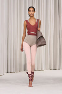 Model walks a fashion runway in a burgundy wrap top, taupe high-waisted bottoms, pale pink leggings, and ribbon-tied sandals, carrying a large taupe bag.