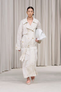 Model walking on a runway in a crinkled beige trench coat, belt tied, carrying a white bag, wearing pale pink heels.