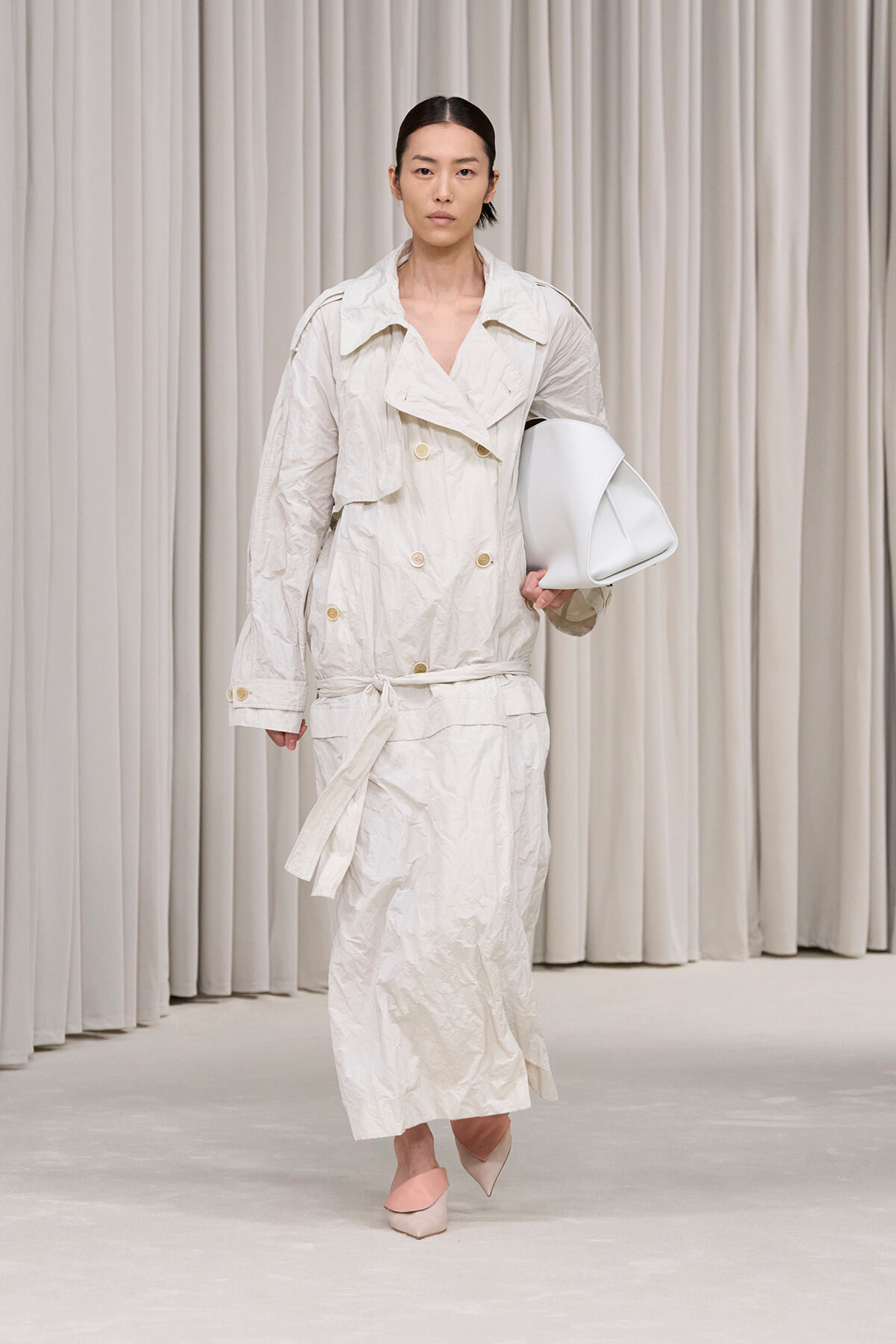 Model walking on a runway in a crinkled beige trench coat, belt tied, carrying a white bag, wearing pale pink heels.