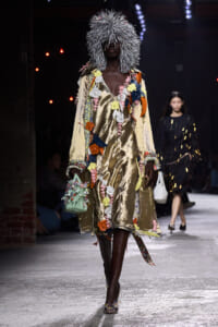 Model walking a runway in a shiny gold dress covered with colorful fabric flowers, wearing a silver spiky wig and carrying a small floral handbag.