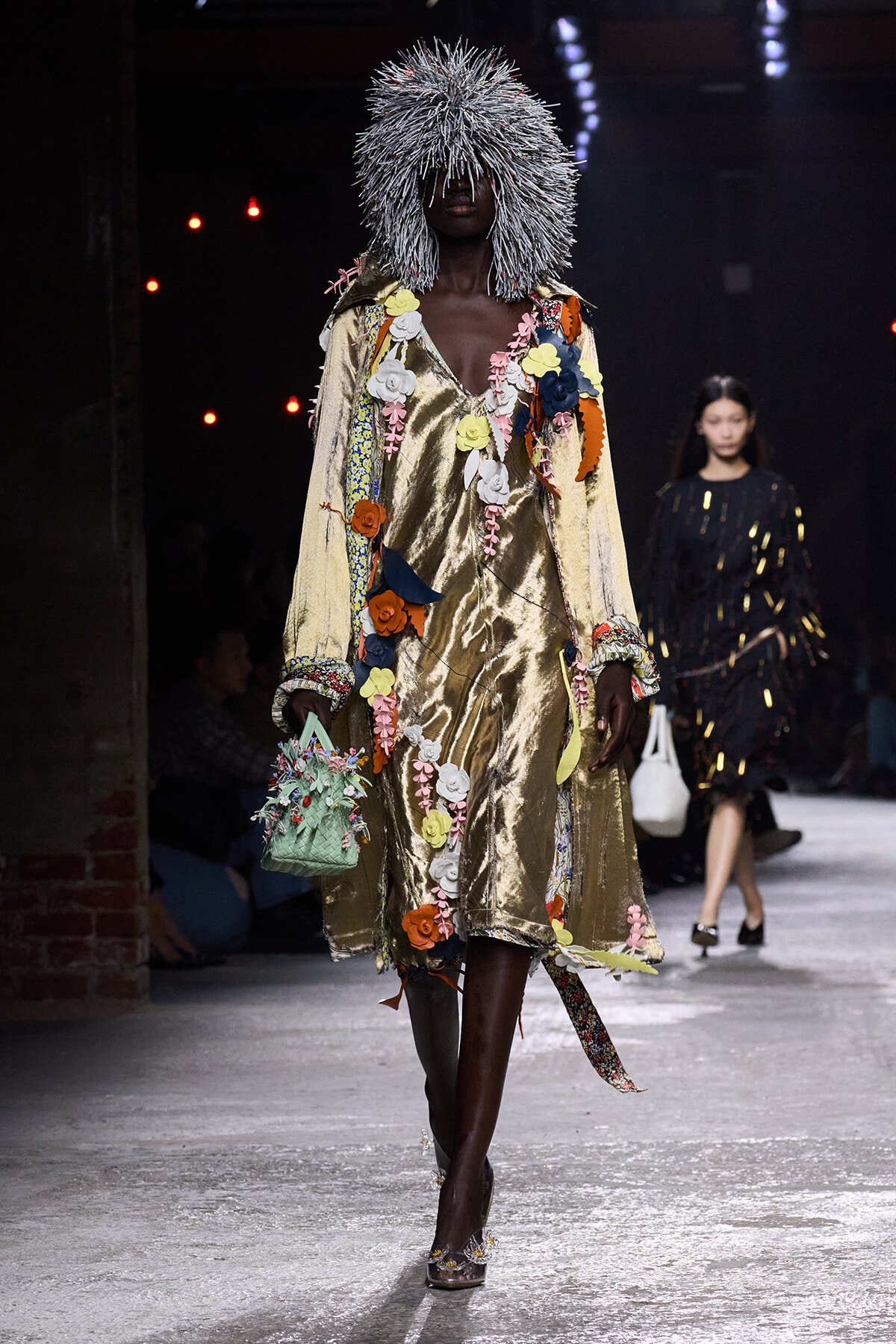 Model walking a runway in a shiny gold dress covered with colorful fabric flowers, wearing a silver spiky wig and carrying a small floral handbag.