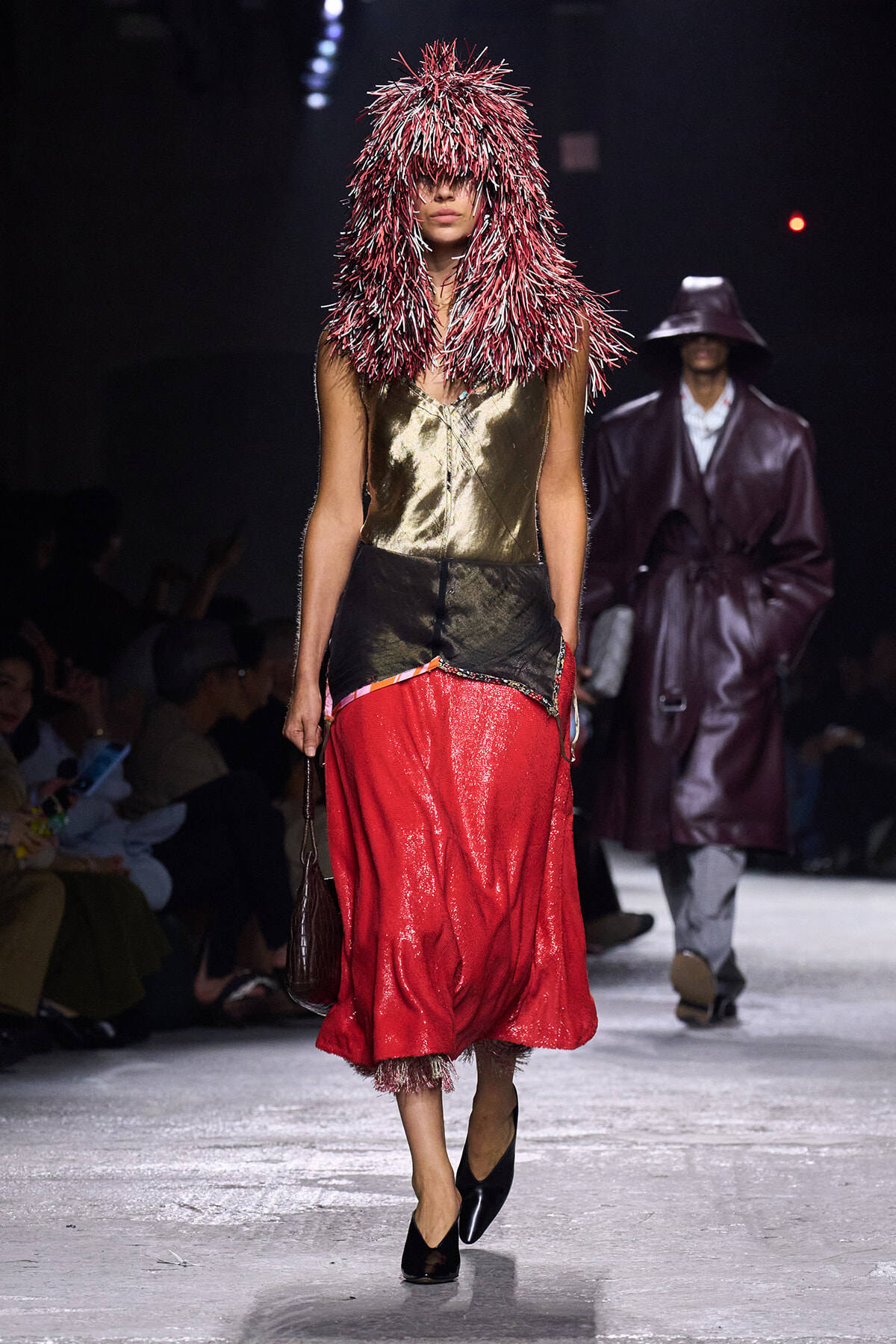 Model walking a fashion show in a metallic gold top and red sequined skirt, wearing a large red-and-white spiky hood that covers the face.