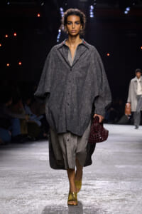 Female model walks the runway in a oversized gray textured coat over layered gray garments, carrying a burgundy woven handbag.