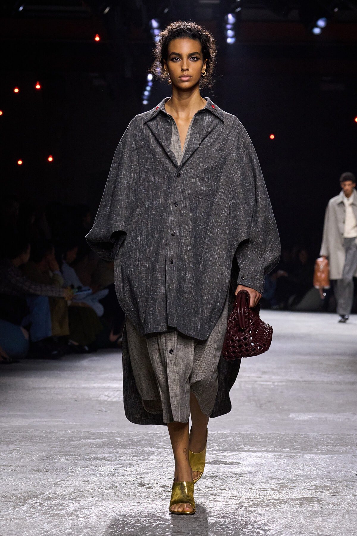 Female model walks the runway in a oversized gray textured coat over layered gray garments, carrying a burgundy woven handbag.