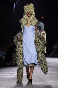 Model on a runway wearing a beige shaggy fur coat with hood over a light blue slip dress.