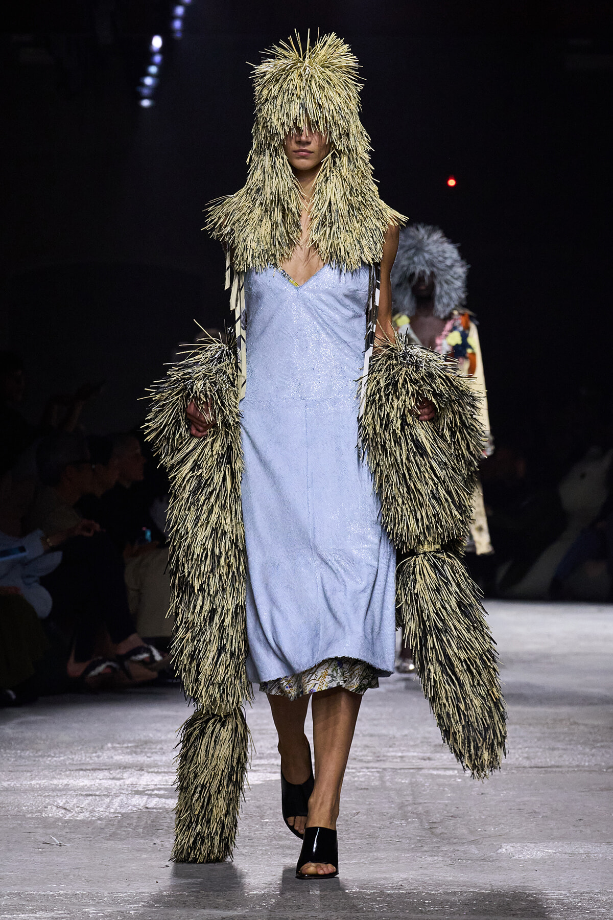Model on a runway wearing a beige shaggy fur coat with hood over a light blue slip dress.