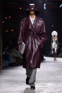 Model on a runway wearing a burgundy leather trench with wide belt and oversized collar, paired with a wide-brim hat and a gray quilted clutch.