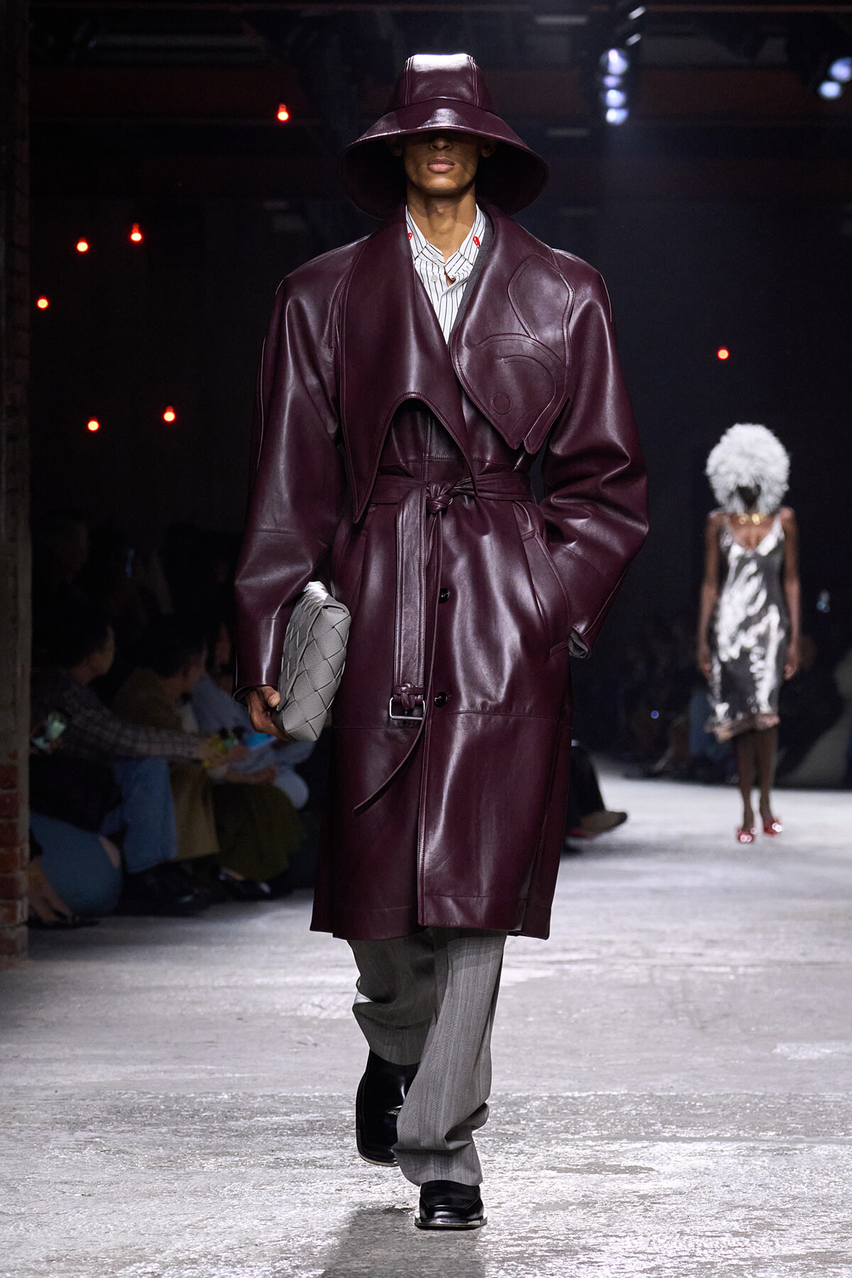 Model on a runway wearing a burgundy leather trench with wide belt and oversized collar, paired with a wide-brim hat and a gray quilted clutch.