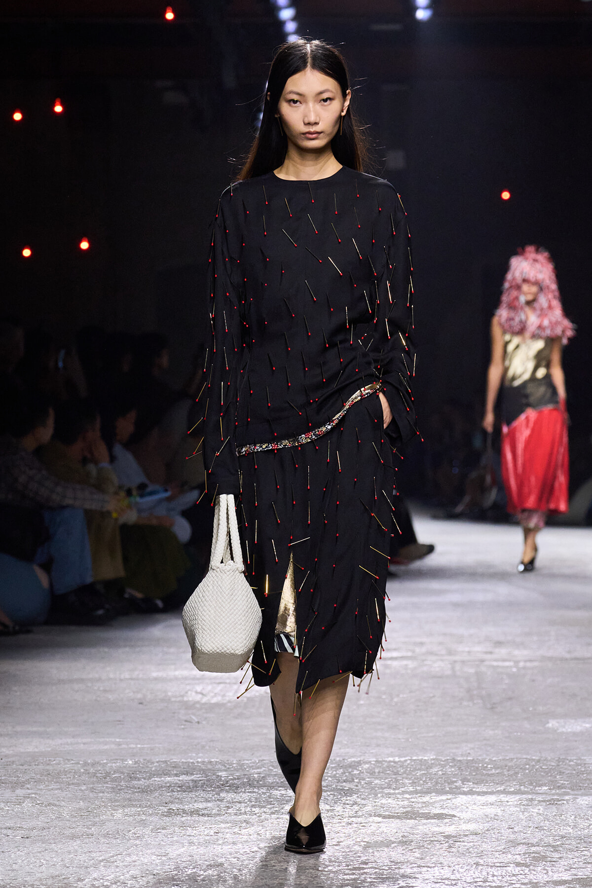 Model walks the runway in a black, pin-studded dress with a white woven bag, minimalist makeup, and black flats.