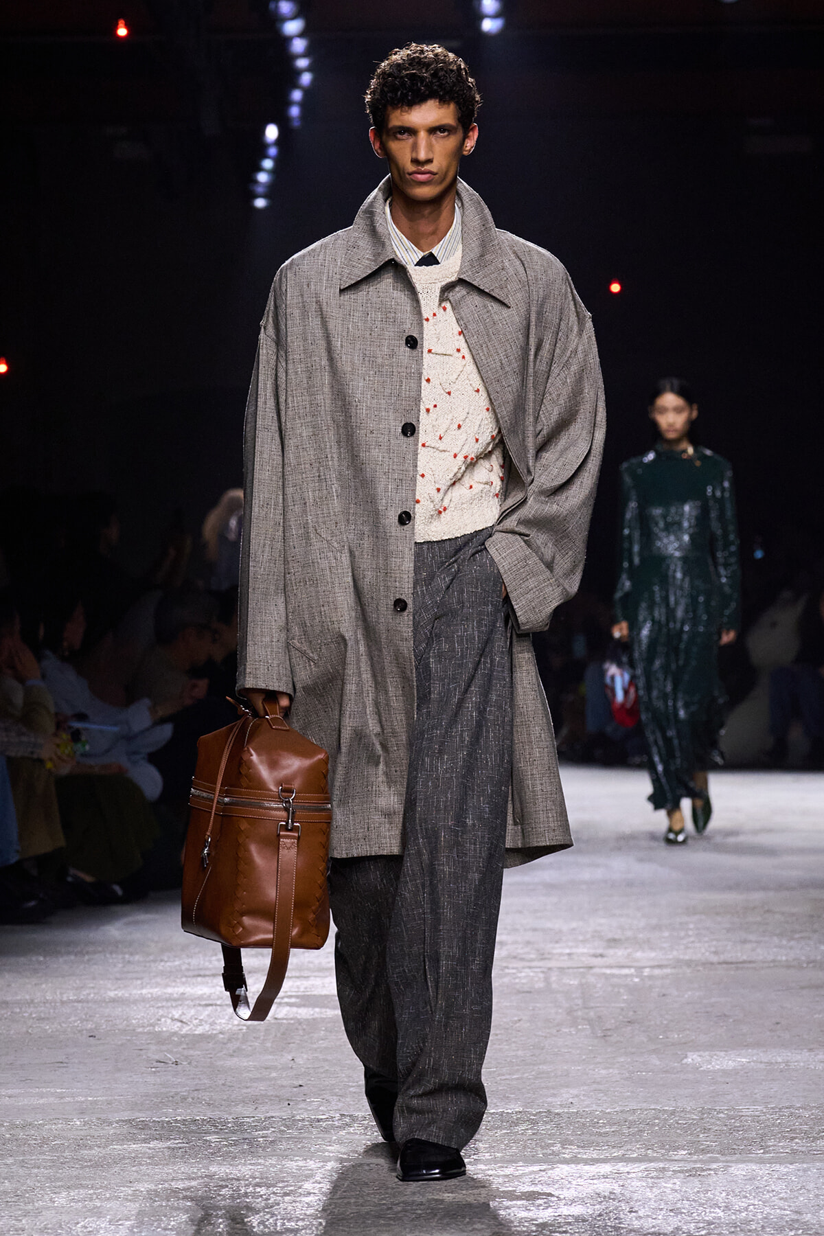 Male model on a runway wearing a oversized gray coat, white knit sweater, dark trousers, and a brown leather backpack on his side.