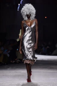 Model walking runway in a metallic silver slip dress with a voluminous white fringe headpiece and red embellished heels, floral underlayer peeking out