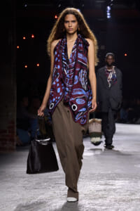 Model walking a fashion runway wearing a burgundy-blue draped top with abstract print and wide brown trousers, carrying a large black tote.