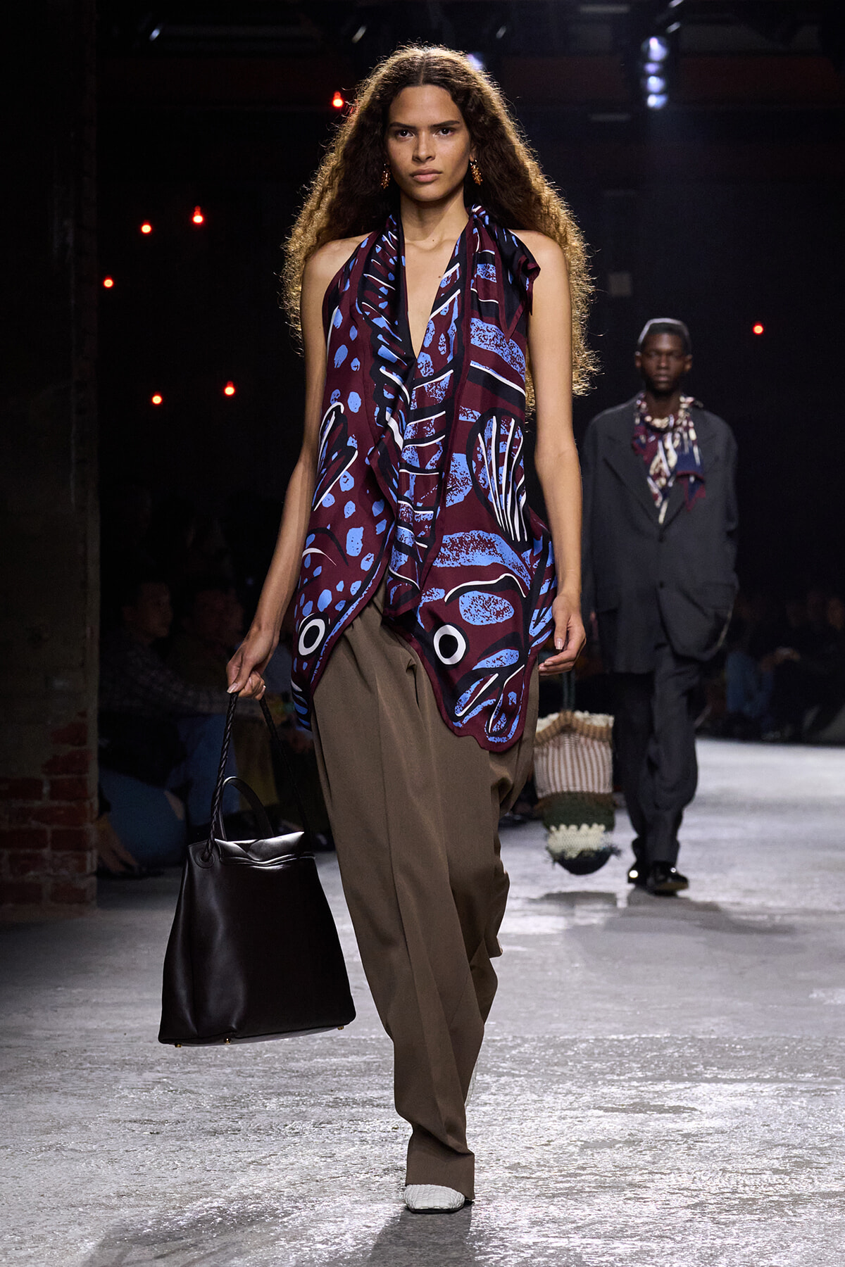Model walking a fashion runway wearing a burgundy-blue draped top with abstract print and wide brown trousers, carrying a large black tote.