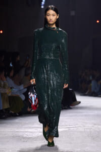 Model walking a fashion show in a dark emerald-green, long-sleeve, sequined dress, carrying a colorful fringed bag.