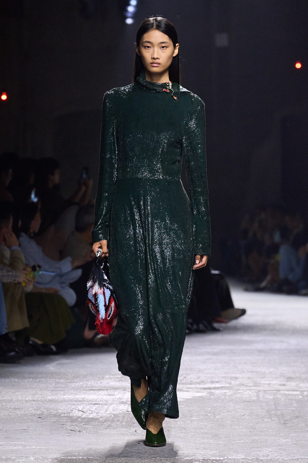 Model walking a fashion show in a dark emerald-green, long-sleeve, sequined dress, carrying a colorful fringed bag.