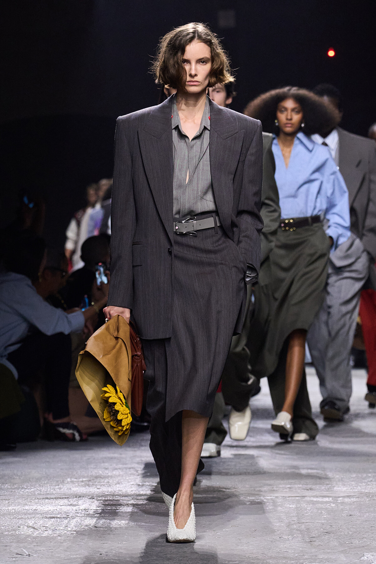 Model walking a fashion runway in a oversized gray pinstripe suit, gray blouse, belt, and white woven heels, carrying a tan hat with sunflowers.