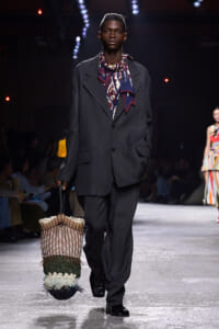 Male model walks a runway in an oversized charcoal suit with a patterned scarf, carrying a large woven tote with fringe.