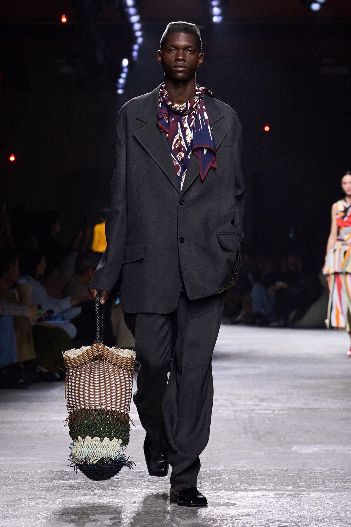 Male model walks a runway in an oversized charcoal suit with a patterned scarf, carrying a large woven tote with fringe.