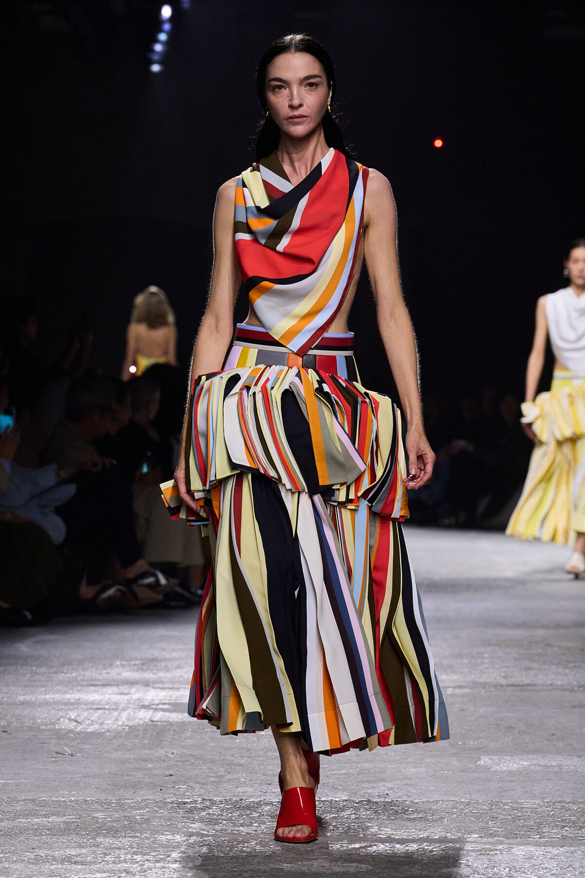 Model on a runway wearing a bold multicolor striped draped top and voluminous striped skirt with red sandals.