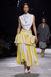 Model walks the runway in a white draped sleeveless top with a yellow-and-white striped, tiered skirt and white sandals.
