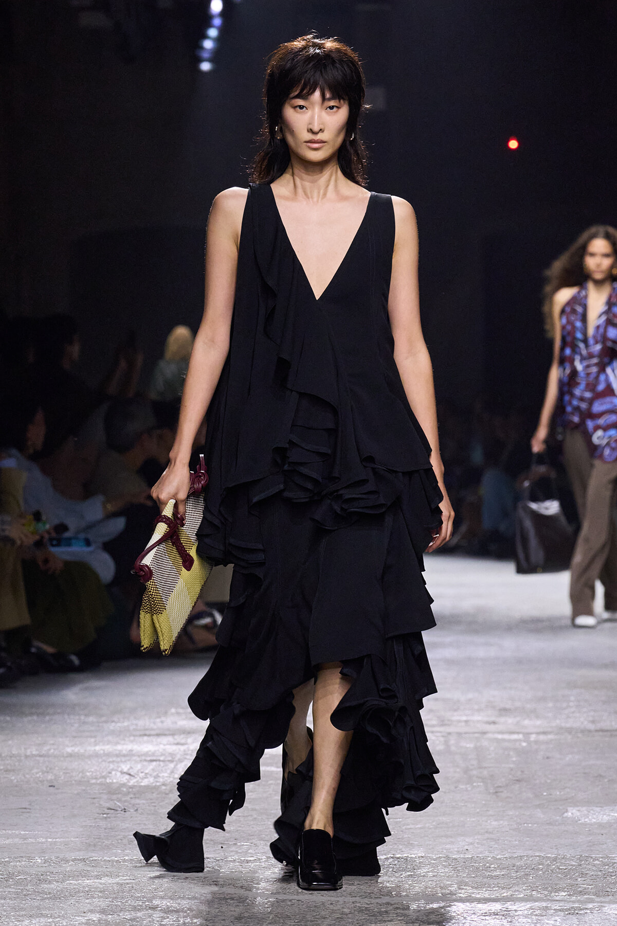 Model walks a runway in a sleeveless black V-neck dress with cascading ruffles, carrying a woven bag.