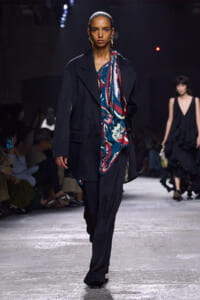 Model walks the runway in an oversized navy blazer over a bold blue-red scarf and loose black trousers. Audience blurred in background.