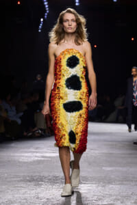 Model walks the runway wearing a strapless knee-length dress made of layered yellow, orange, white, and black floral petals forming a seed-like pattern, holding a small burgundy clutch.