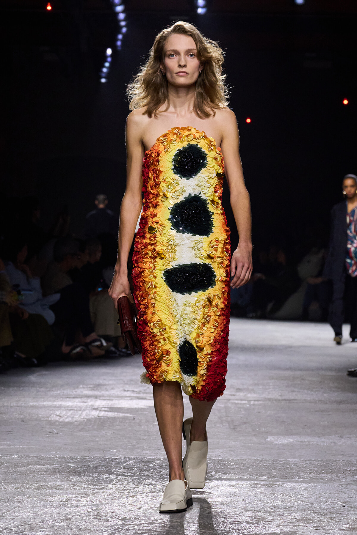 Model walks the runway wearing a strapless knee-length dress made of layered yellow, orange, white, and black floral petals forming a seed-like pattern, holding a small burgundy clutch.