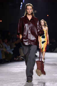 Model walks a fashion show wearing a glossy burgundy short-sleeve shirt with wide collar and grey wide-leg trousers, carrying a woven red-brown handbag.