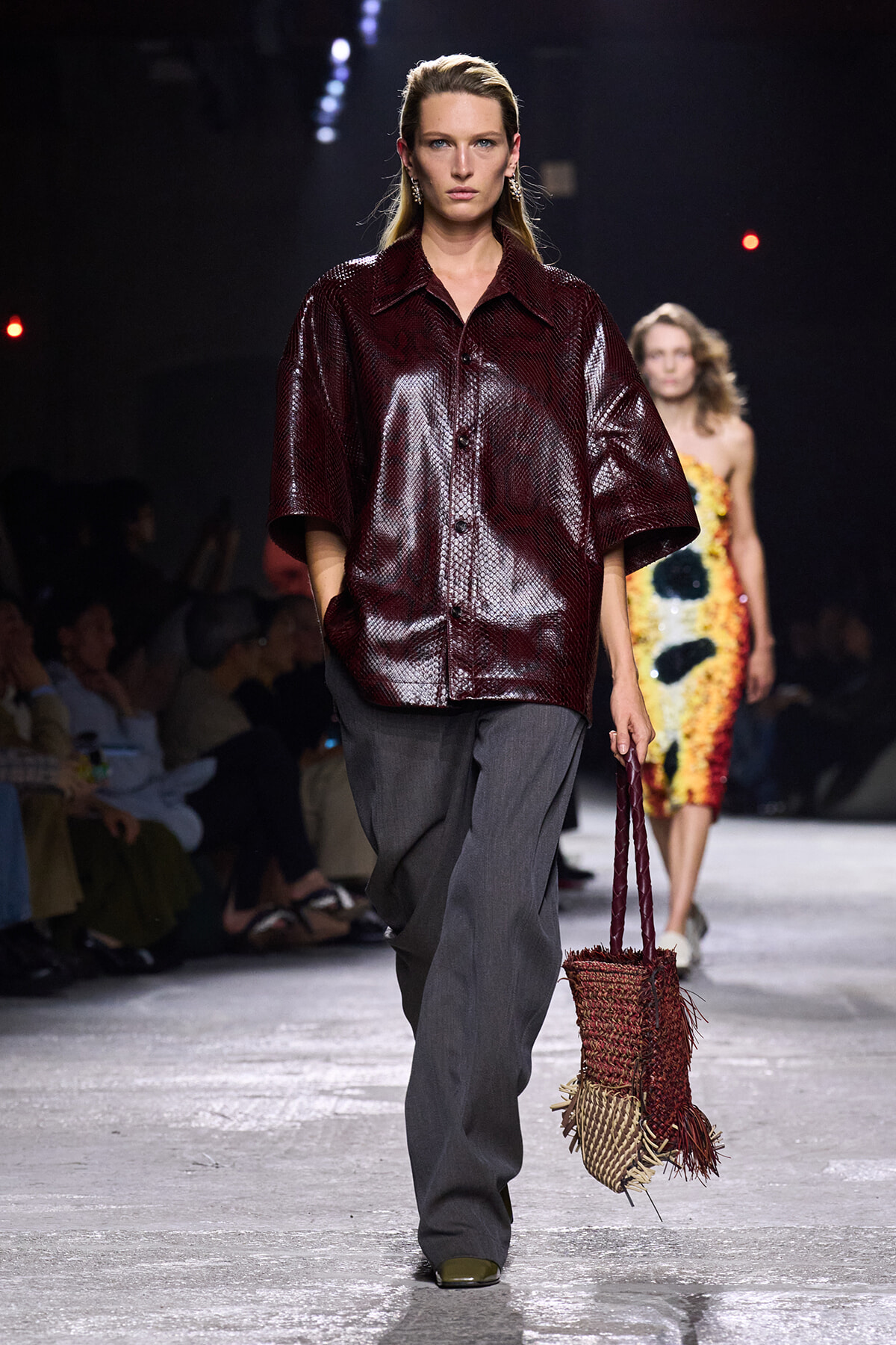 Model walks a fashion show wearing a glossy burgundy short-sleeve shirt with wide collar and grey wide-leg trousers, carrying a woven red-brown handbag.