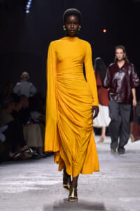Model on a runway wearing a mustard-yellow draped midi dress with one cape-style sleeve and green earrings.