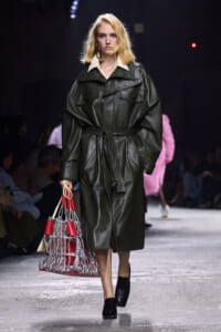 Model on a runway wearing an oversized dark green leather trench coat, belted at the waist, with a red-and-silver lattice handbag in hand.