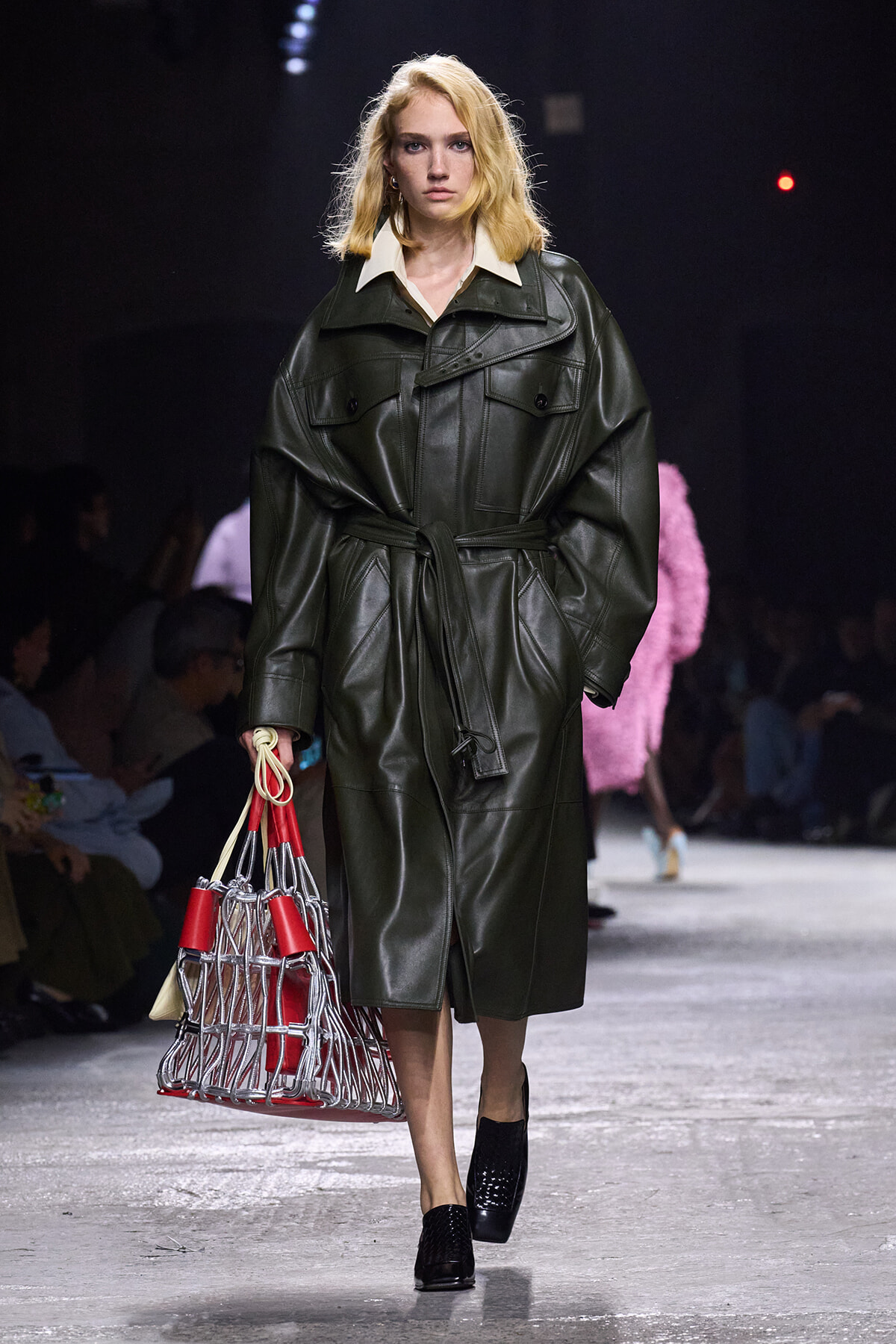 Model on a runway wearing an oversized dark green leather trench coat, belted at the waist, with a red-and-silver lattice handbag in hand.