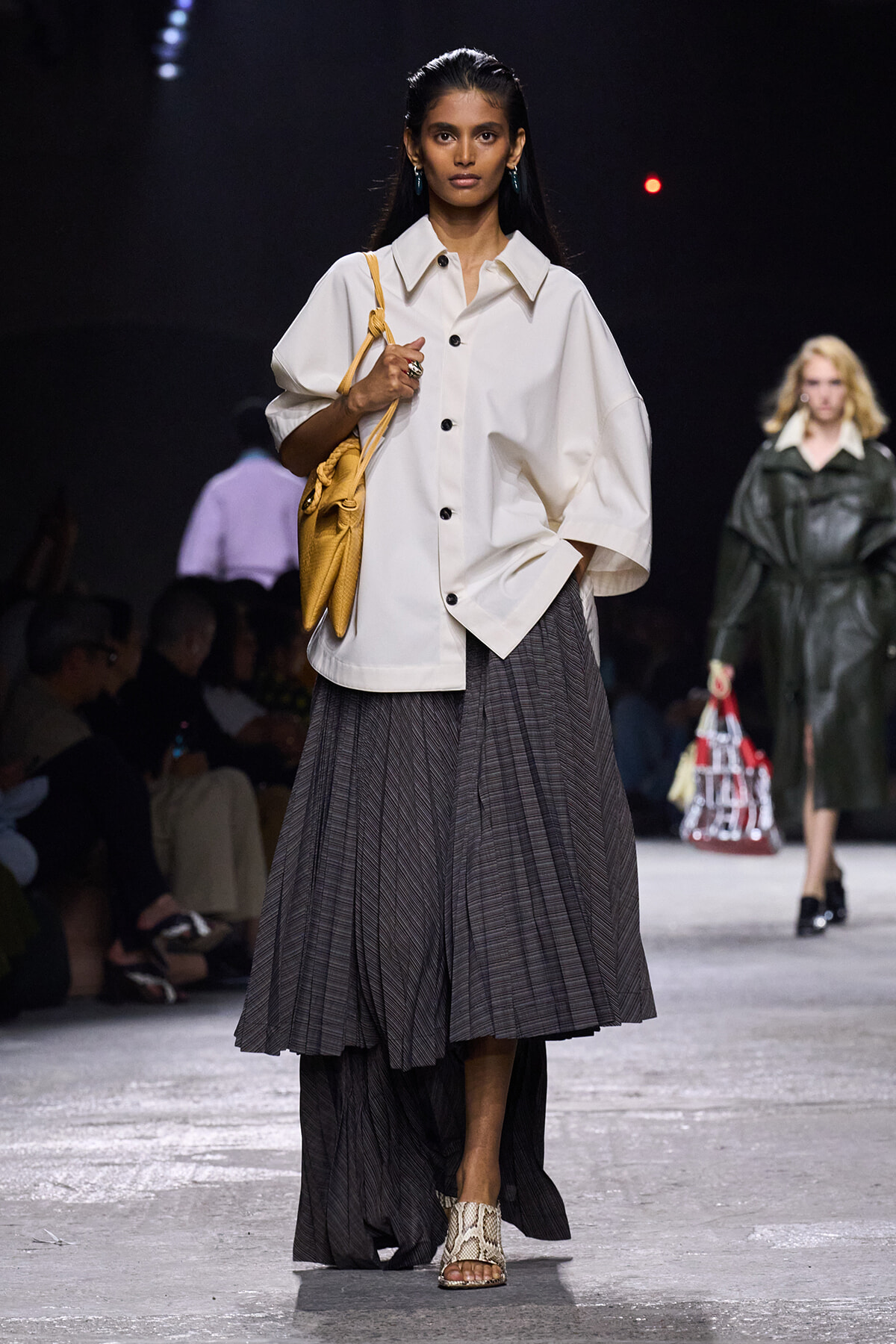 Model walking a runway in an oversized white button-front blouse, grey pleated midi skirt, snake-print sandals, and a yellow shoulder bag.
