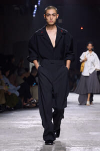 Male runway model in an all-black, oversized shirt with a deep V neck and wide-leg trousers, hands in pockets as he walks the catwalk.