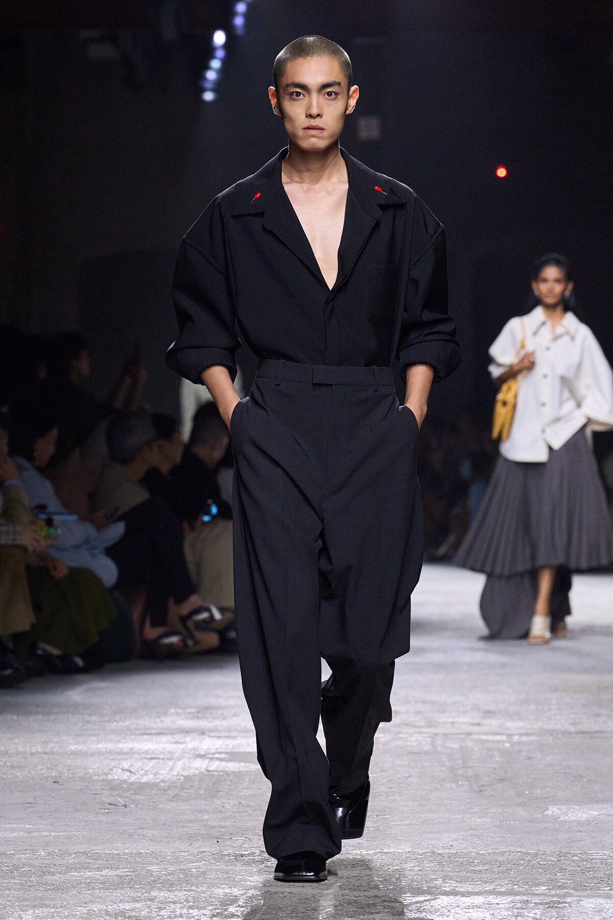 Male runway model in an all-black, oversized shirt with a deep V neck and wide-leg trousers, hands in pockets as he walks the catwalk.