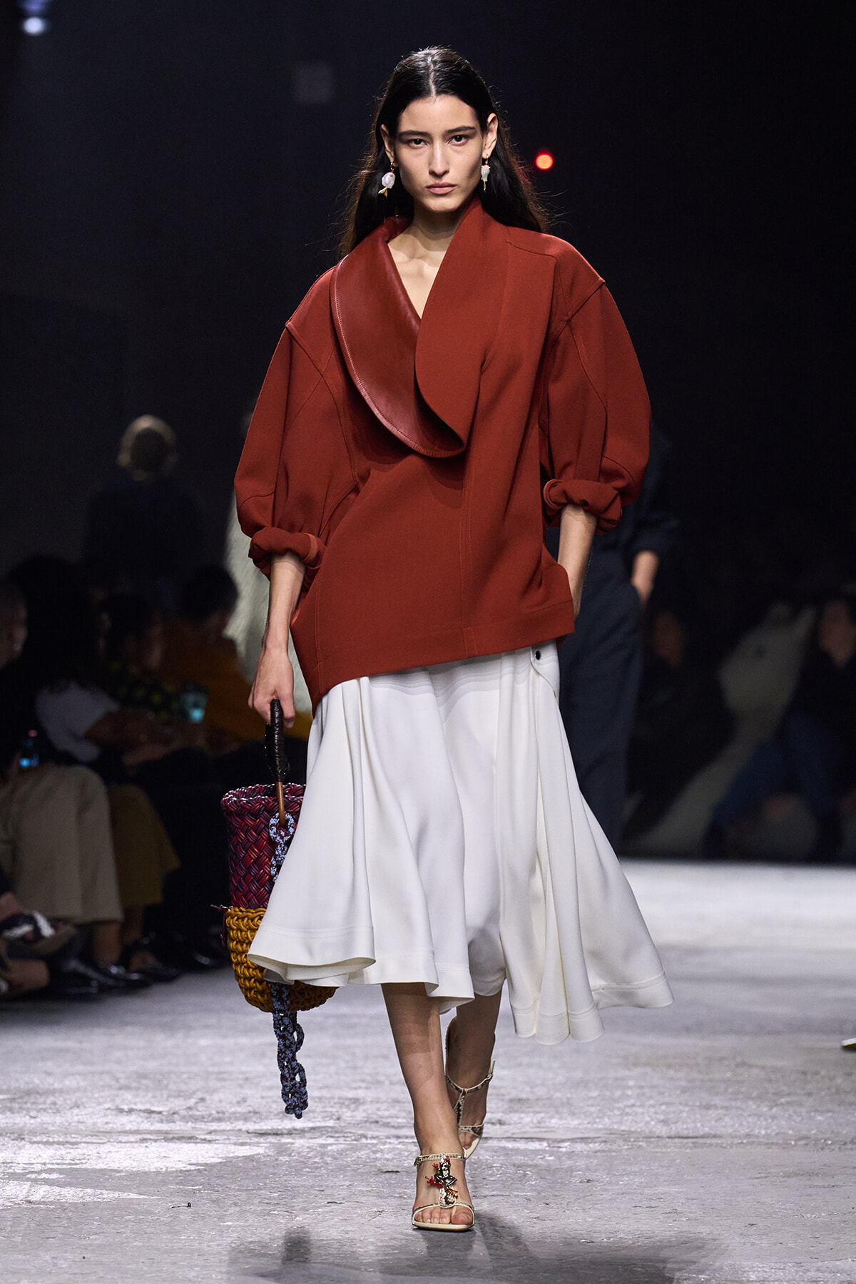 Model walks the runway in an oversized red coat with a large draped collar, paired with a white flowing skirt and a woven handbag.
