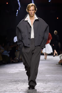 Male model walks a fashion show runway in an oversized charcoal gray jacket, white shirt, and wide gray trousers with his hands in pockets.