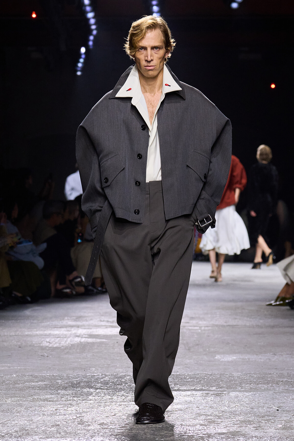 Male model walks a fashion show runway in an oversized charcoal gray jacket, white shirt, and wide gray trousers with his hands in pockets.