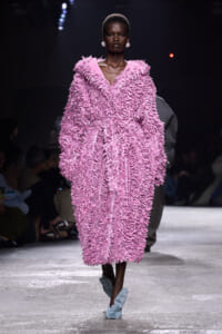 Model walks the runway in a voluminous pink shaggy coat with textured fringe and pale blue fuzzy slippers.
