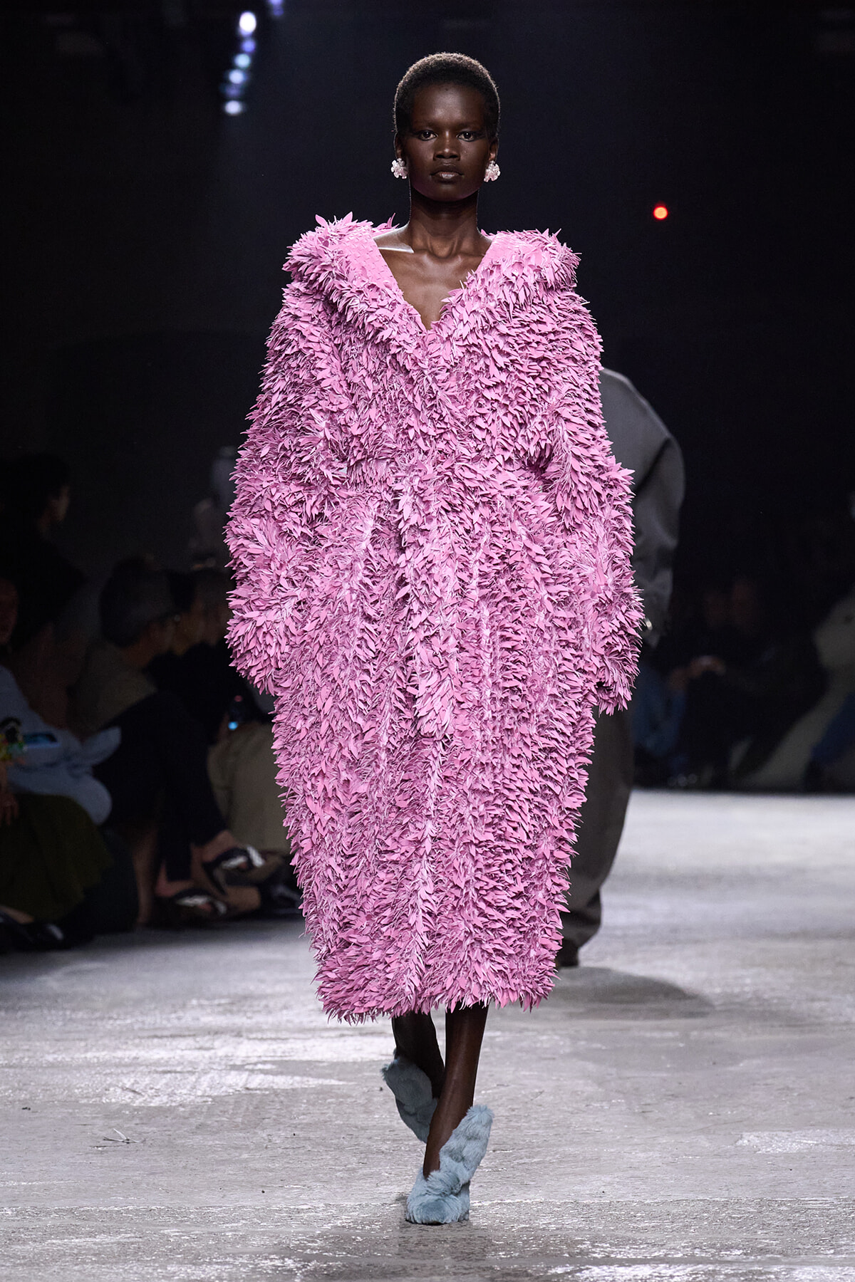 Model walks the runway in a voluminous pink shaggy coat with textured fringe and pale blue fuzzy slippers.