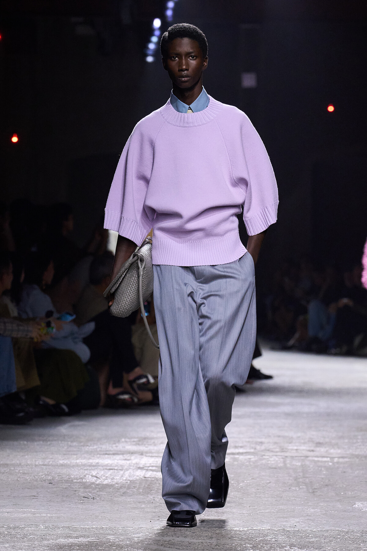 Model walks the runway in a lavender oversized knit sweater layered over a light blue collared shirt, with loose gray trousers and a textured gray handbag.
