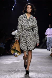 Female model walking a runway in a black-and-white striped belted coat, carrying a brown paper bag with flowers inside.] repeated?